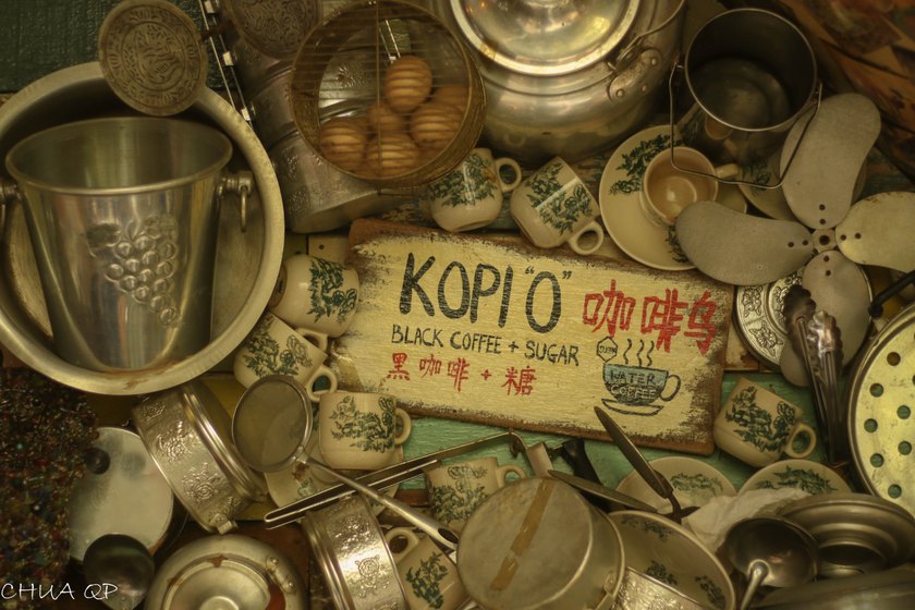 A hand-painted kopi sign surrounded by vintage cups, kettles and metal kitchen utensils